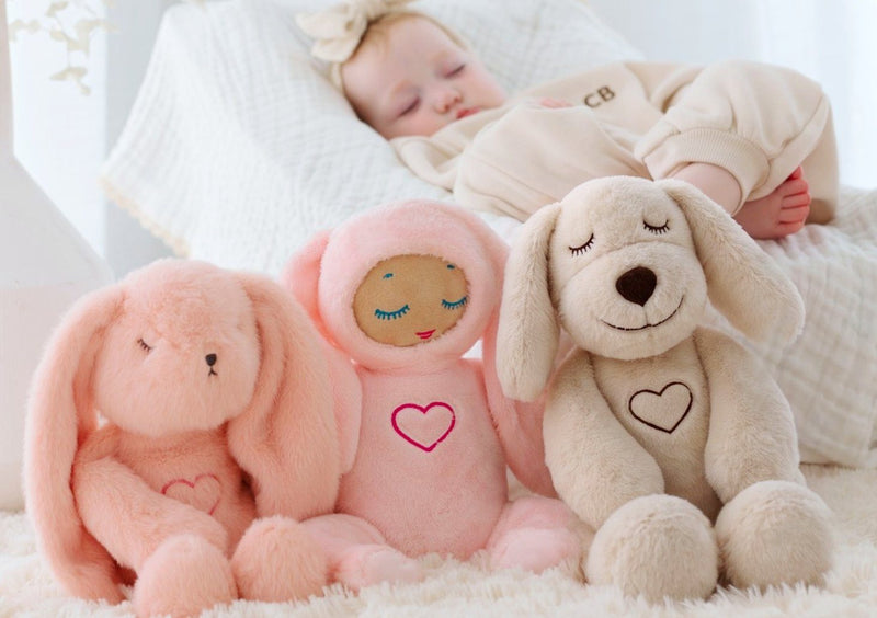 A baby asleep behind a Lulla Bunny, Lulla Doll and Lulla Pup